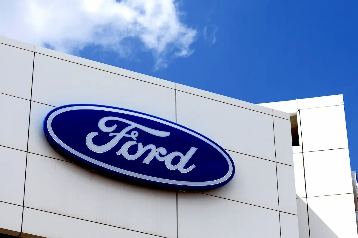 Blue Ford oval logo mounted on the white paneled exterior of a building against a blue sky with scattered clouds.